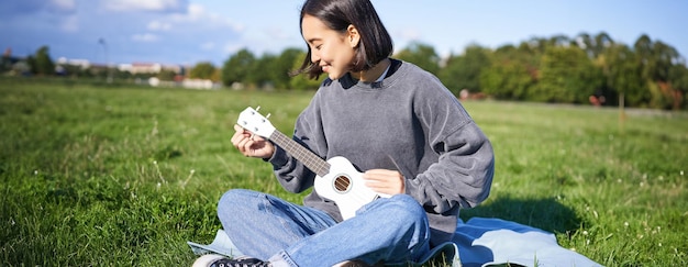 Foto gratuito ragazza sorridente asiatica che sintonizza la sua chitarra ukulele cantando e suonando nel parco seduta su una coperta al sole