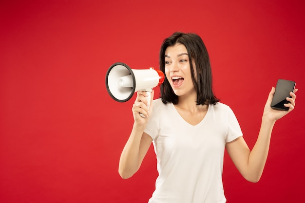 Foto gratuito ritratto a mezzo busto della giovane donna caucasica su sfondo rosso studio. bello modello femminile in camicia. concetto di emozioni umane, espressione facciale, vendite. tenendo il telefono e chiamando a bocca aperta.