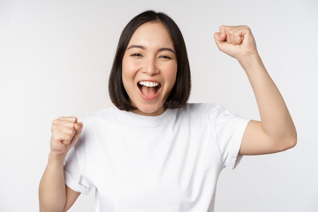 Foto gratuito ritratto di donna asiatica entusiasta che vince celebrando e trionfando alzando le mani per raggiungere l'obiettivo o il successo in piedi su sfondo bianco