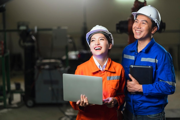 Foto gratuito ritratto di tecnico maschio femmina ingegnere asiatico in uniforme di sicurezza in piedi e girarsi per guardare la fotocamera e ridere sorridere con allegro e sicuro di sé sullo sfondo del posto di lavoro della fabbrica di macchinari