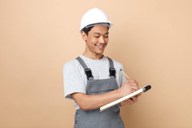 Foto gratuito sorridente giovane operaio edile che indossa il casco di sicurezza e scrittura uniforme con la matita sul blocco note