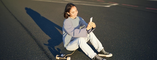 Foto gratuito una ragazza asiatica felice si siede sullo skateboard, si fa un selfie con il longboard, fa facce carine in una giornata di sole all'aperto.