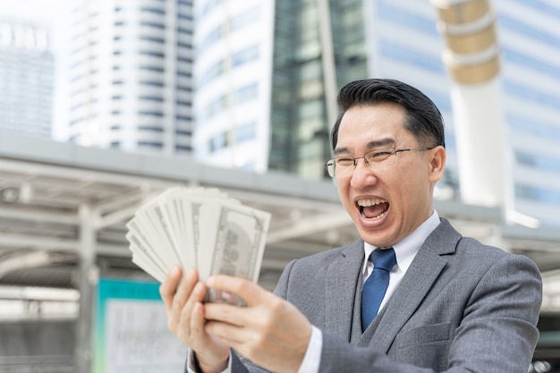 Foto gratuito uomo d'affari asiatico faccia felice che tiene soldi banconote in dollari usa sul quartiere degli affari urbano