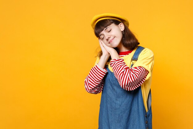 Foto adolescente ragazza rilassata in berretto francese, prendisole in denim tenendo gli occhi chiusi, dormendo con le mani vicino al viso isolato su sfondo giallo. persone sincere emozioni, concetto di stile di vita. mock up spazio di copia.