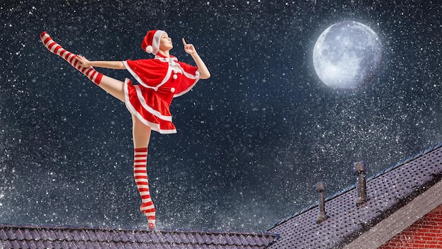 Ballerina ballerina con un regalo tra le mani in un costume da Babbo Natale sul tetto di una casa la notte di Natale sullo sfondo del cielo stellato