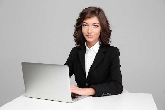 Bella giovane donna d'affari riccia sicura che si siede e lavora usando il laptop