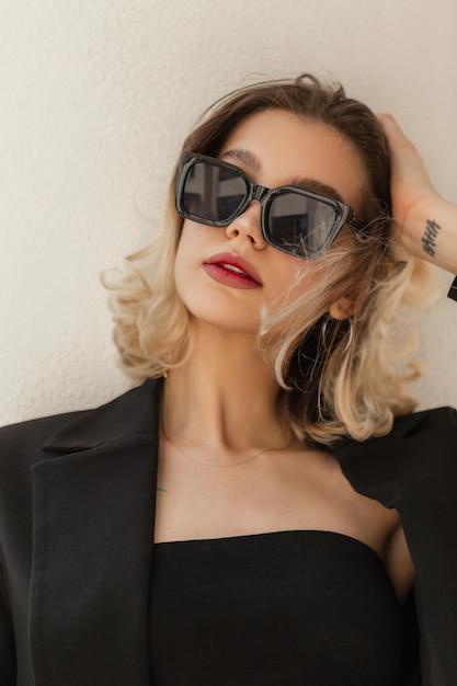 Bella giovane donna elegante modello hipster in occhiali da sole neri alla moda con blazer nero da lavoro e top in piedi vicino al muro bianco sulla strada