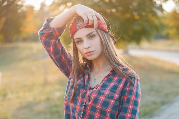 Bella giovane hipster con un berretto di baseball rosso in una camicia alla moda nel parco al tramonto