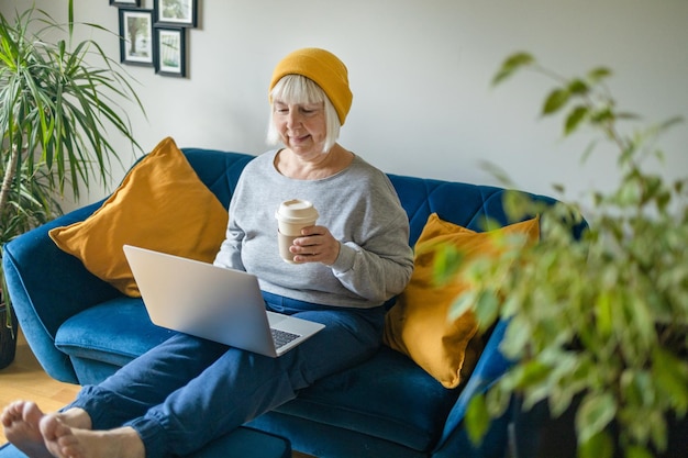 Donna di mezza età degli anni '50 in possesso di caffè caldo che lavora al computer portatile seduto a casa in ufficio