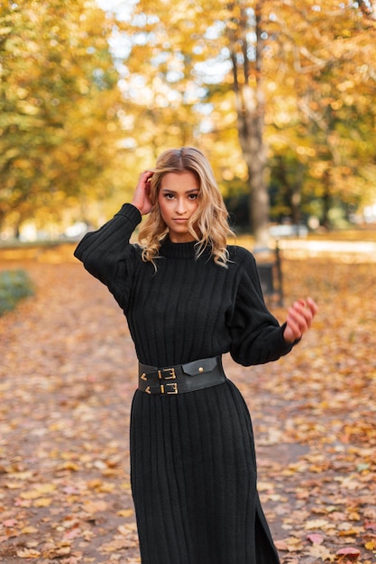 Elegante bella giovane donna bionda in un vestito a maglia nera di moda cammina nel parco d'autunno