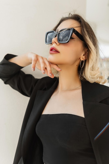 Elegante e bella giovane donna modello di moda con occhiali da sole alla moda in abiti neri alla moda con un blazer e top in posa fuori da un edificio bianco vintage in una giornata di sole