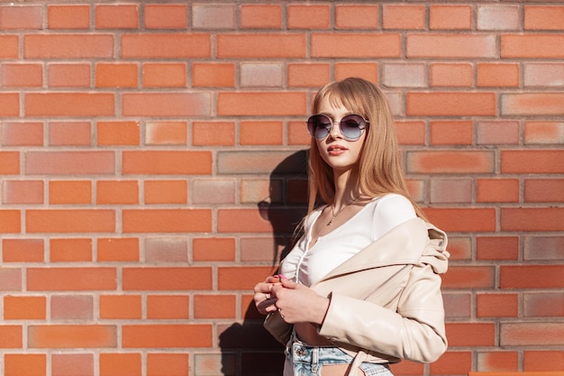 Elegante e bella ragazza modello con occhiali da sole alla moda e vestiti alla moda si trova vicino a un muro di mattoni vintage in città Moda e bellezza in stile femminile urbano