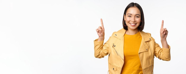 Entusiasta ragazza asiatica che punta il dito verso l'alto mostrando pubblicità in cima sorridente felice dimostrando offerta promozionale o banner in piedi su sfondo bianco