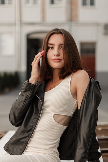 Felice giovane bella ragazza caucasica in un abito bianco con una giacca di pelle alla moda si siede sulla strada e guarda la telecamera