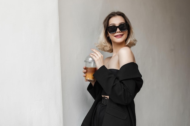 Felice giovane elegante modello di donna sorridente alla moda in occhiali neri alla moda con giacca e pantaloni di moda nera superiore beve caffè freddo e cammina per strada vicino a un muro bianco