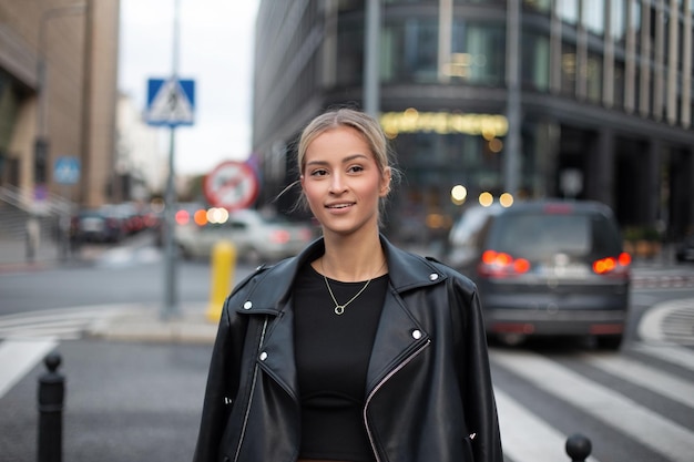 Felice giovane modella urbana in abito di moda con giacca nera di pelle che cammina in città Signora piuttosto chic sullo sfondo urbano