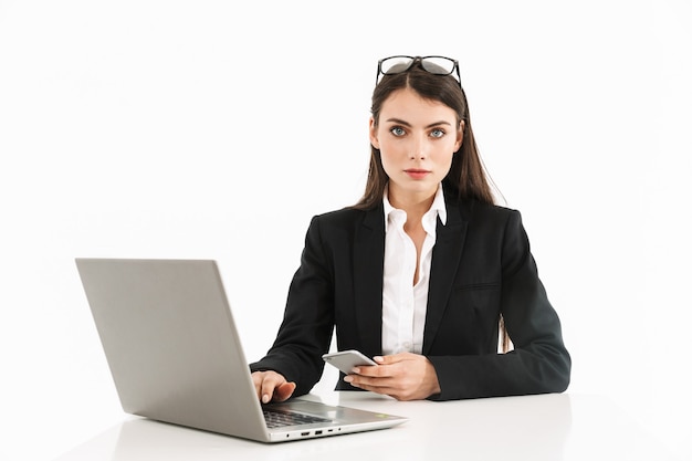 Foto di una donna d'affari seria lavoratrice vestita con abiti formali seduta alla scrivania e che lavora al computer portatile in ufficio isolato su un muro bianco