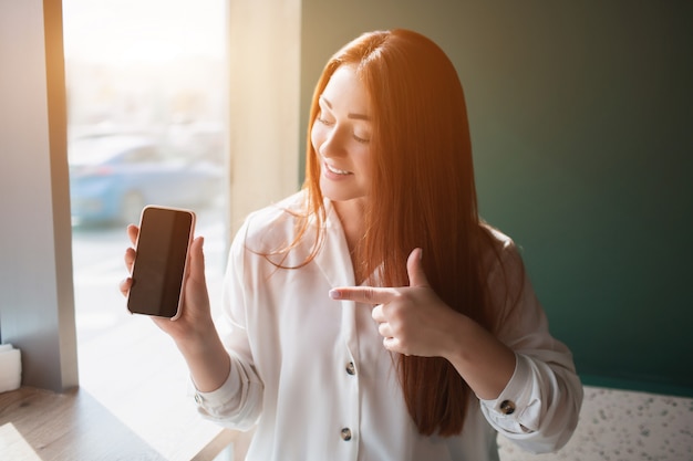 Giovane donna dai capelli rossi che si siede in un caffè e che tiene un telefono. Il modello femminile punta con un dito indice sullo smartphone.