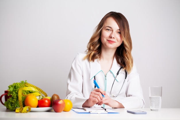 Giovane medico dietista presso la stanza di consulto al tavolo con frutta e verdura fresca, lavorando su un piano di dieta