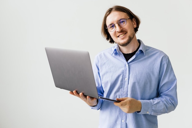 Giovane uomo bello sicuro in computer portatile della tenuta della camicia e sorridere mentre stando contro il fondo bianco