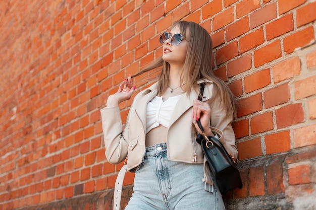 Il ritratto di strada delle donne di una bella ragazza carina ed elegante in abito alla moda con occhiali da sole alla moda giacca di pelle jeans a vita alta e borsa si trova vicino a un muro di mattoni rossi in città