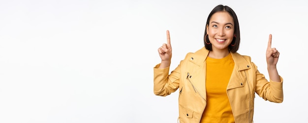 Immagine di una donna bruna asiatica sorridente che punta il dito verso l'alto mostrando pubblicità con la faccia felice in posa su sfondo bianco