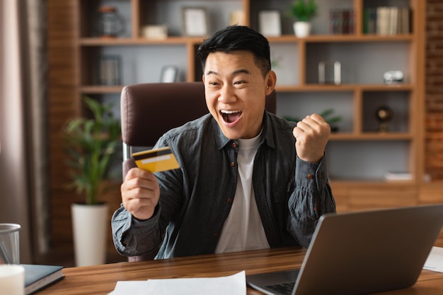 Foto l'uomo d'affari giapponese adulto eccitato sorpreso soddisfatto con la carta di credito della tenuta della bocca aperta fa il gesto di vittoria