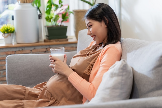 La donna incinta asiatica tiene un bicchiere di latte per la sua salute e il bambino con il sorriso e seduto sul divano, sentendosi felice e rilassato. Concetto di donne incinte.