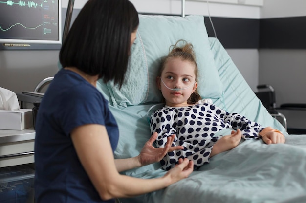 Madre premurosa che parla con la figlia malata ricoverata che riposa nel letto del paziente del reparto pediatrico dell'ospedale mentre è in trattamento farmacologico. Genitore premuroso seduto accanto alla bambina malata mentre la conforta.