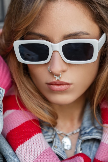 Modello alla moda giovane bella donna con occhiali da sole bianchi con piercing al naso in abiti denim urbani di moda in città Ritratto femminile grazioso della ragazza rossa