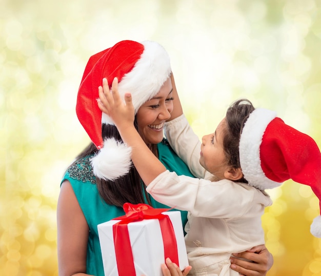 Natale, vacanze, celebrazione, famiglia e concetto di persone - madre felice e bambina con cappelli di Babbo Natale con confezione regalo su sfondo a luci gialle