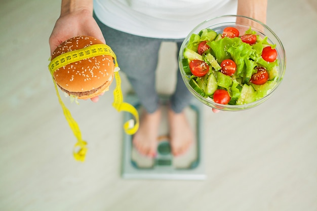 Peso corporeo di misurazione della donna sull'hamburger e sull'insalata della tenuta della bilancia.