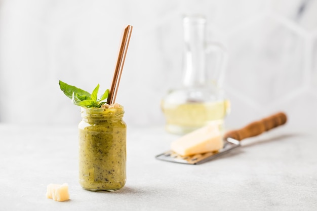 Foto pesto fatto in casa in un vasetto con una foglia di basilico una bottiglia di olio d'oliva una fetta di parmigiano e un cucchiaio cucina italiana