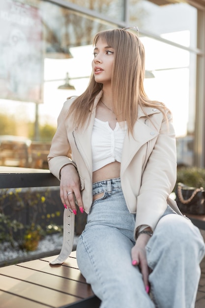 Piuttosto giovane donna in giacca rock in pelle con top bianco e jeans blu vintage si siede su una panca di legno sulla strada Abito casual urbano stile femminile alla moda