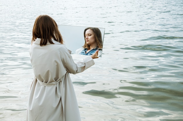 Primo piano della donna che sta su una riva in vestito blu che tiene uno specchio con la sua riflessione su