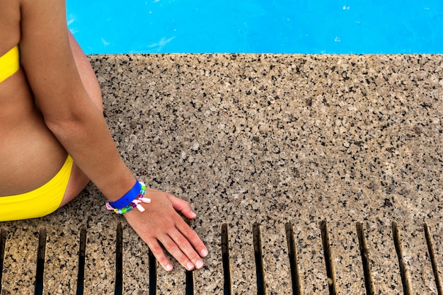 Primo piano della giovane donna che indossa il costume da bagno giallo del bikini che riposa vicino alla piscina con chiara acqua blu il giorno soleggiato di estate.