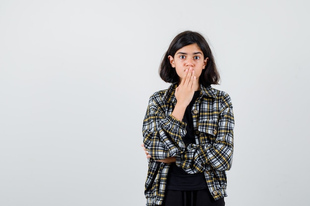 Ragazza teenager che tiene la mano sulla bocca in camicia casual e sembra scioccata, vista frontale.