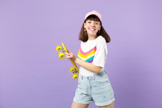 Foto ritratto di sorridente gioiosa ragazza adolescente in abiti vivaci che tiene skateboard giallo isolato su sfondo viola muro pastello in studio. persone sincere emozioni, concetto di stile di vita. mock up spazio di copia.