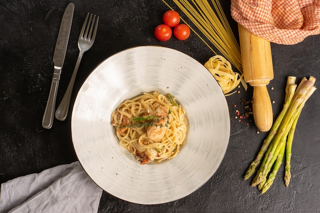 Foto spaghetti con gamberi e asparagi. piatto caldo decorato con pasta, asparagi, pomodorini, un mattarello e posate su una tavola nera. vista dall'alto.