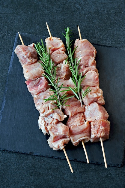 Spiedini crudi con rosmarino e spezie su una tavola di pietra nera.