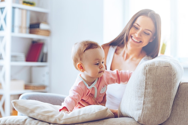 Strisciando sul divano. Piccola neonata che striscia sul divano e si appoggia sul cuscino mentre sua madre è seduta sullo sfondo