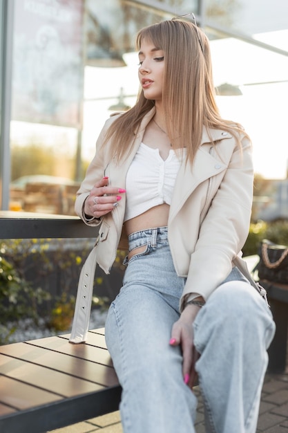 Trendy bella giovane moda hipster ragazza con i capelli in abiti rock alla moda alla moda con un top giacca di pelle e jeans blu vintage si siede su una panca di legno vicino a un caffè al tramonto