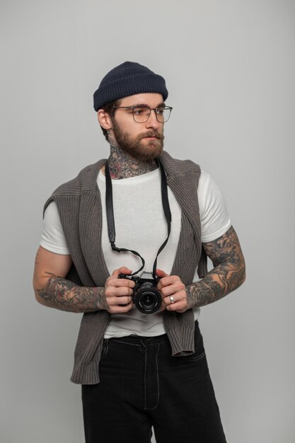 Un uomo elegante di moda, fotografo professionista con un cappello a maglia e un maglione, tiene una fotocamera e sta nello studio.