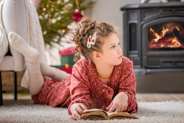 Una bambina carina legge un libro a casa in un interno vissuto vicino a un albero di Natale sullo sfondo di un caminetto acceso