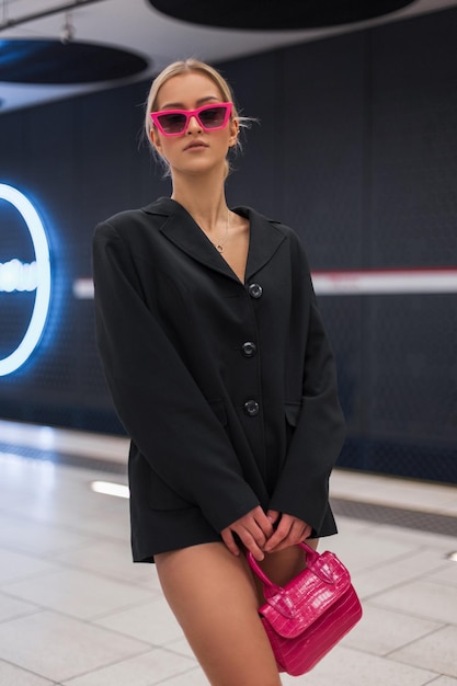 Una bella donna alla moda cool e glamour con occhiali da sole rosa in un blazer nero con una borsa rosa elegante sta e posa in una moderna stazione della metropolitana bianca