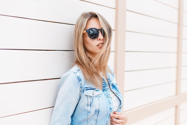 Una bella ragazza adolescente alla moda con occhiali da sole in stile vintage in una giacca denim alla moda e un top in pizzo si trova vicino a un muro di legno