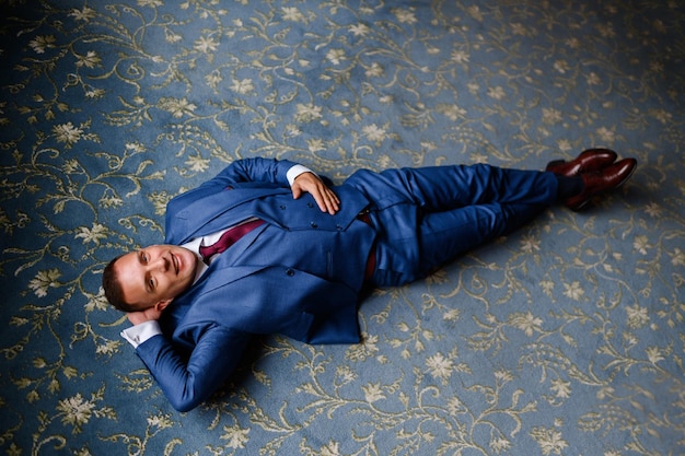 Acima vista do homem feliz vestido de terno elegante e gravata vermelha, deitado no tapete azul com impressão de flores, mantendo a mão atrás da cabeça posando e olhando para a câmera