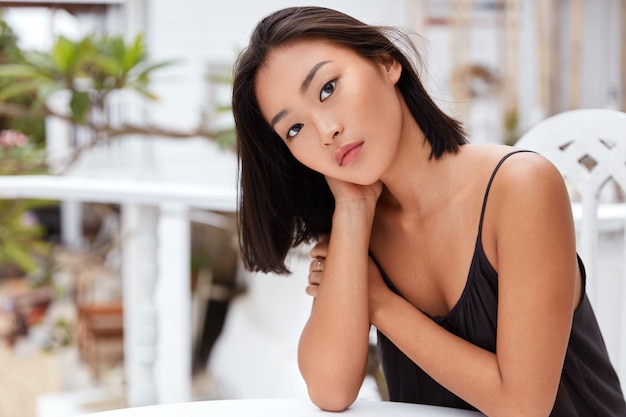 Foto grátis adorável mulher asiática séria tem expressão séria, posa sozinha em um café, sente-se entediada em um país desconhecido, vestida casualmente, tem boa recreação.