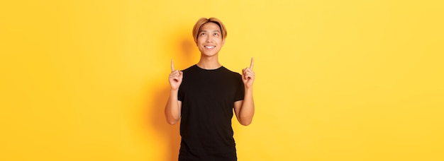 Foto grátis bonito homem asiático sorridente em camiseta preta apontando os dedos para cima olhando para o banner satisfeito amarelo de volta