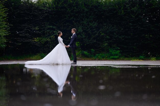 Casal de jovens noivas amorosas de mãos dadas em pé perto do lago olhando um ao outro Mulher noiva linda em vestido inchado com marido homem de terno elegante Celebração do dia do casamento em família